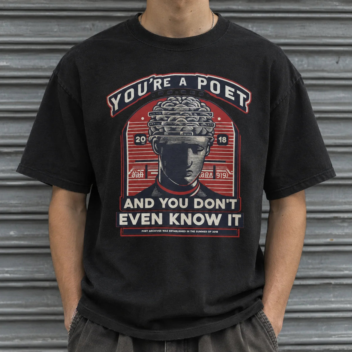 A person wearing a black graphic t-shirt with a red and white design featuring a classical bust illustration and the text 'YOU'RE A POET AND YOU DON'T EVEN KNOW IT', posed in front of a gray metal shutter.