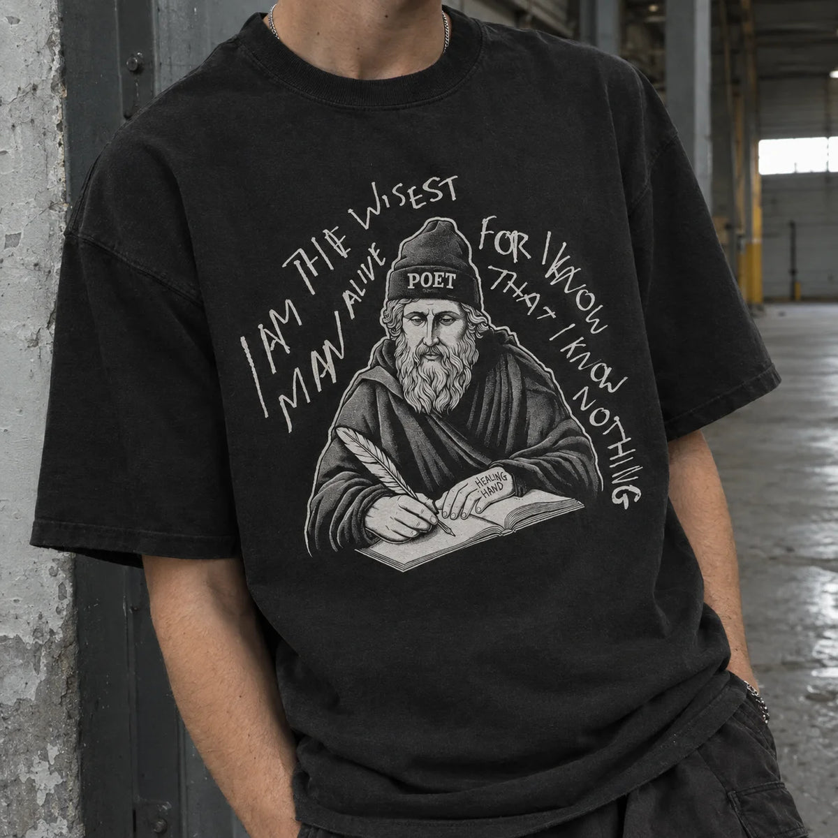 A person wearing a black graphic t-shirt in an industrial warehouse setting. The t-shirt features a white line art illustration of a bearded poet wearing a beanie labeled 'POET' and writing in a book, surrounded by the text 'I AM THE WISEST MAN I'VE EVER KNOW THAT I KNOW NOTHING'.