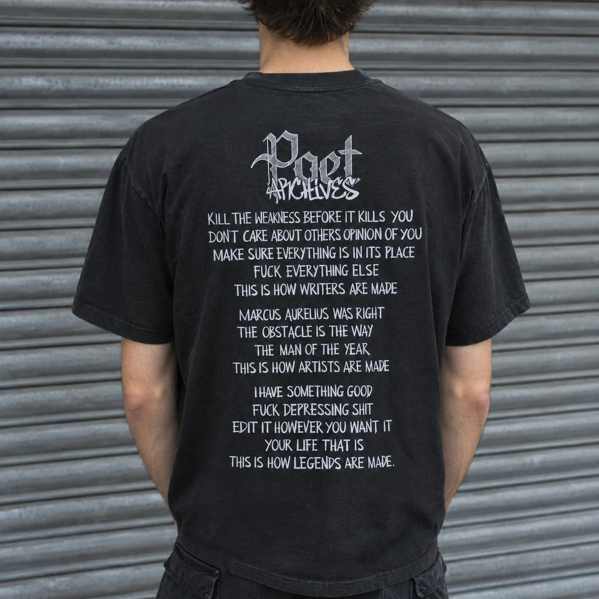 A person viewed from behind wearing a black graphic T-shirt with white text reading 'Poet Archivist' and a long quote, standing in front of a gray metal shutter.
