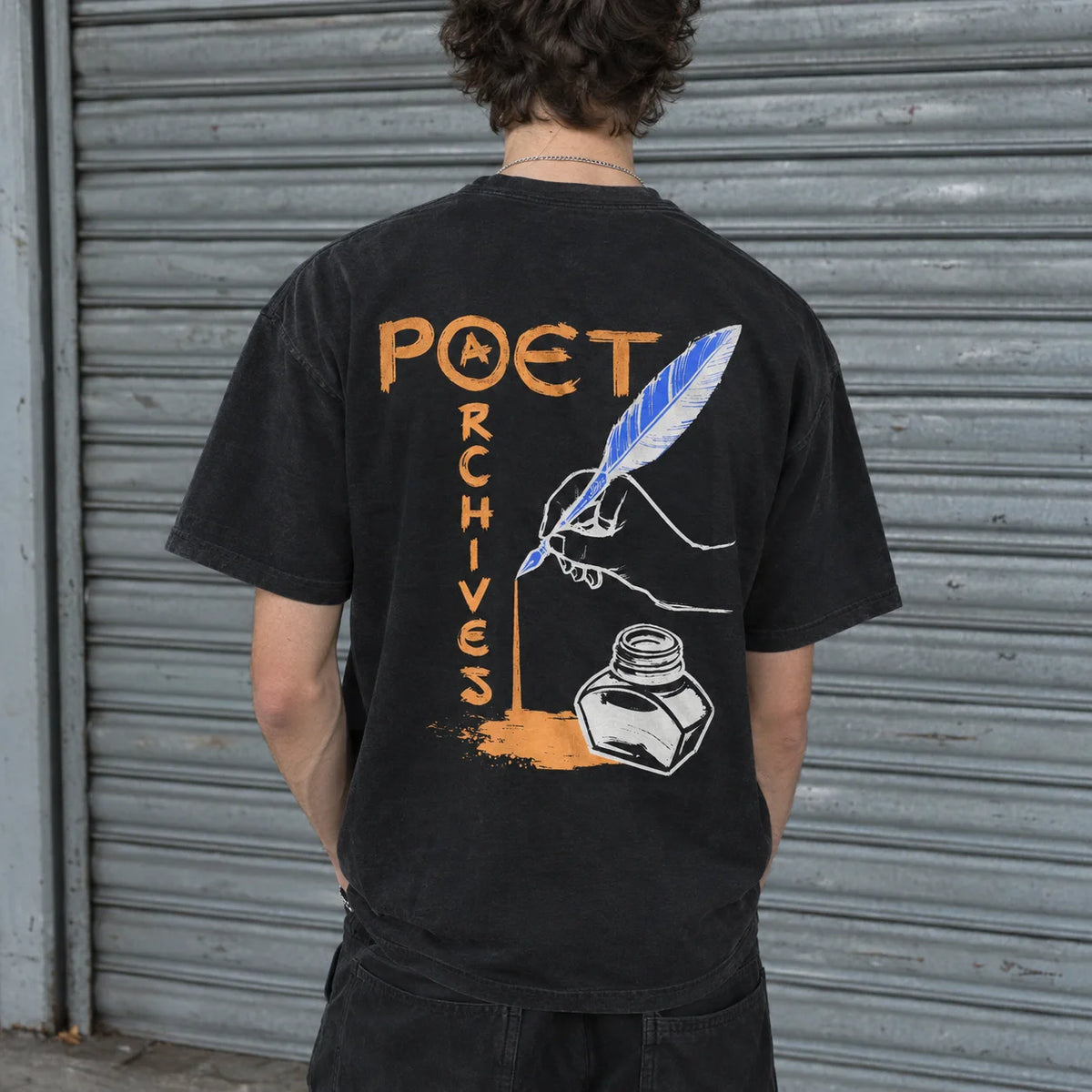 A person viewed from behind wearing a black graphic T-shirt in front of a gray metal roll-up door. The shirt features a large back print with the words “POET ARCHIVES” in orange, a white and blue feather quill, and an ink bottle illustration.