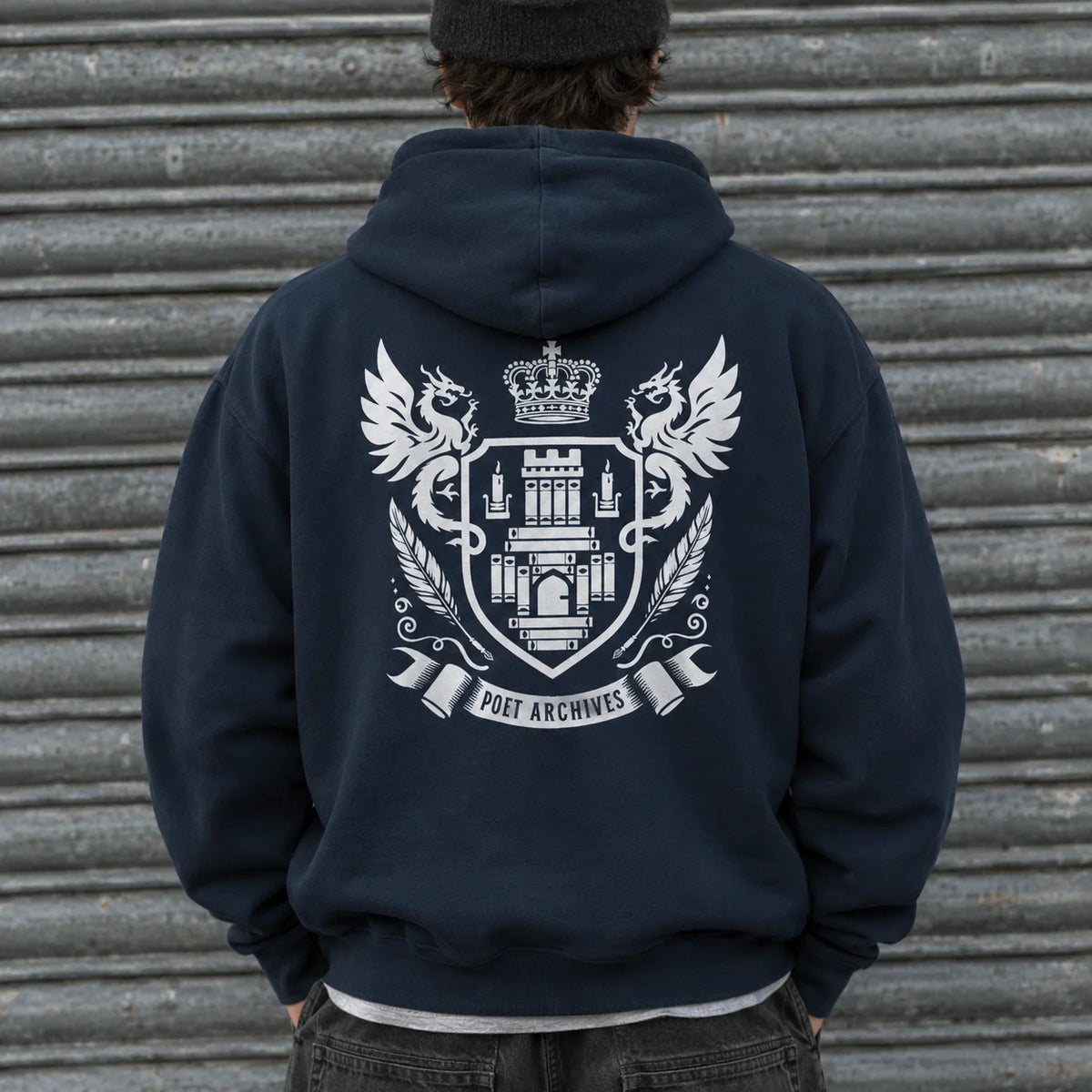 A person viewed from behind wearing a navy blue hoodie with a large white crest graphic on the back, standing in front of a corrugated metal wall.