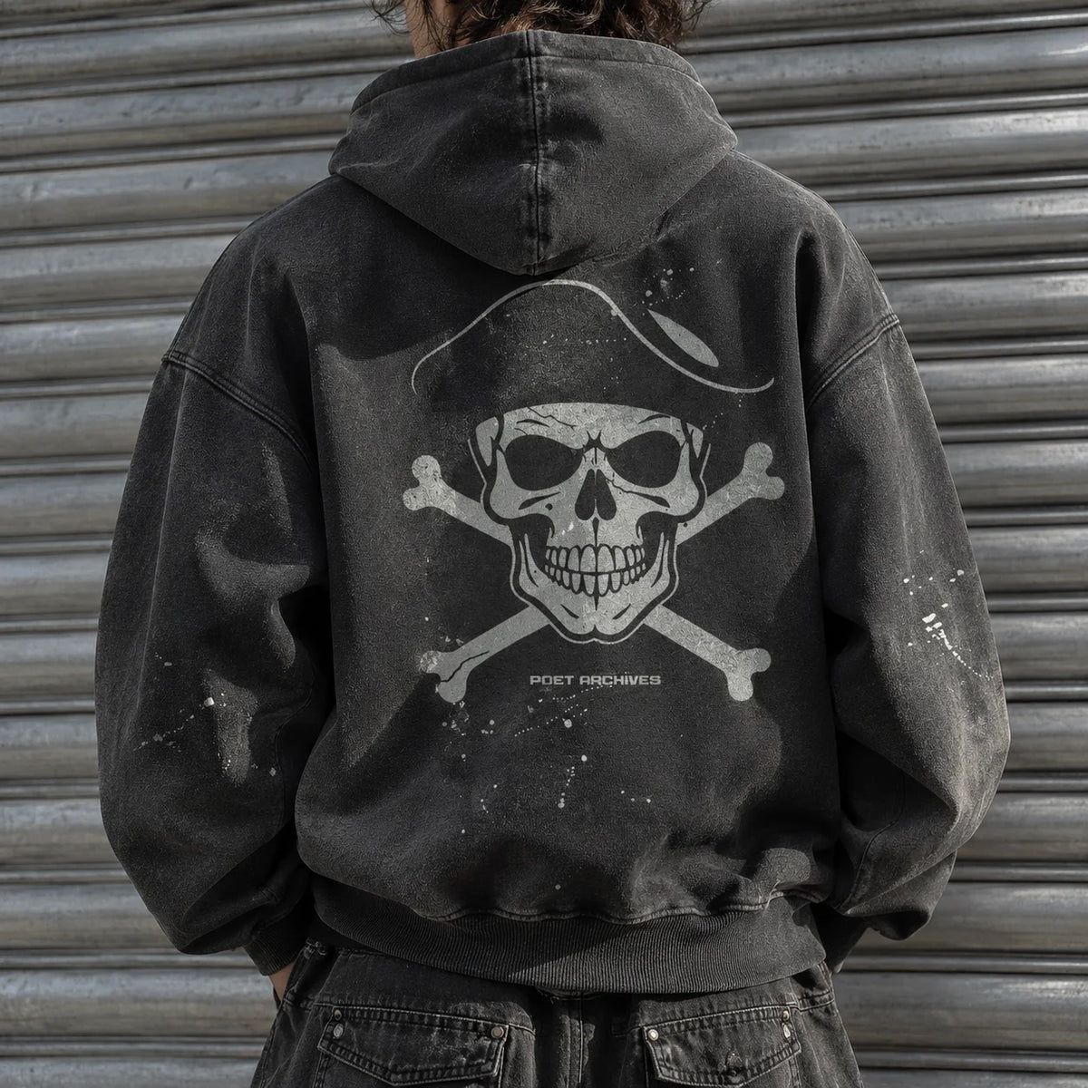 A person viewed from the back wearing a faded black hooded sweatshirt with a large distressed white skull and crossbones graphic on the back, standing in front of a gray metal shutter.