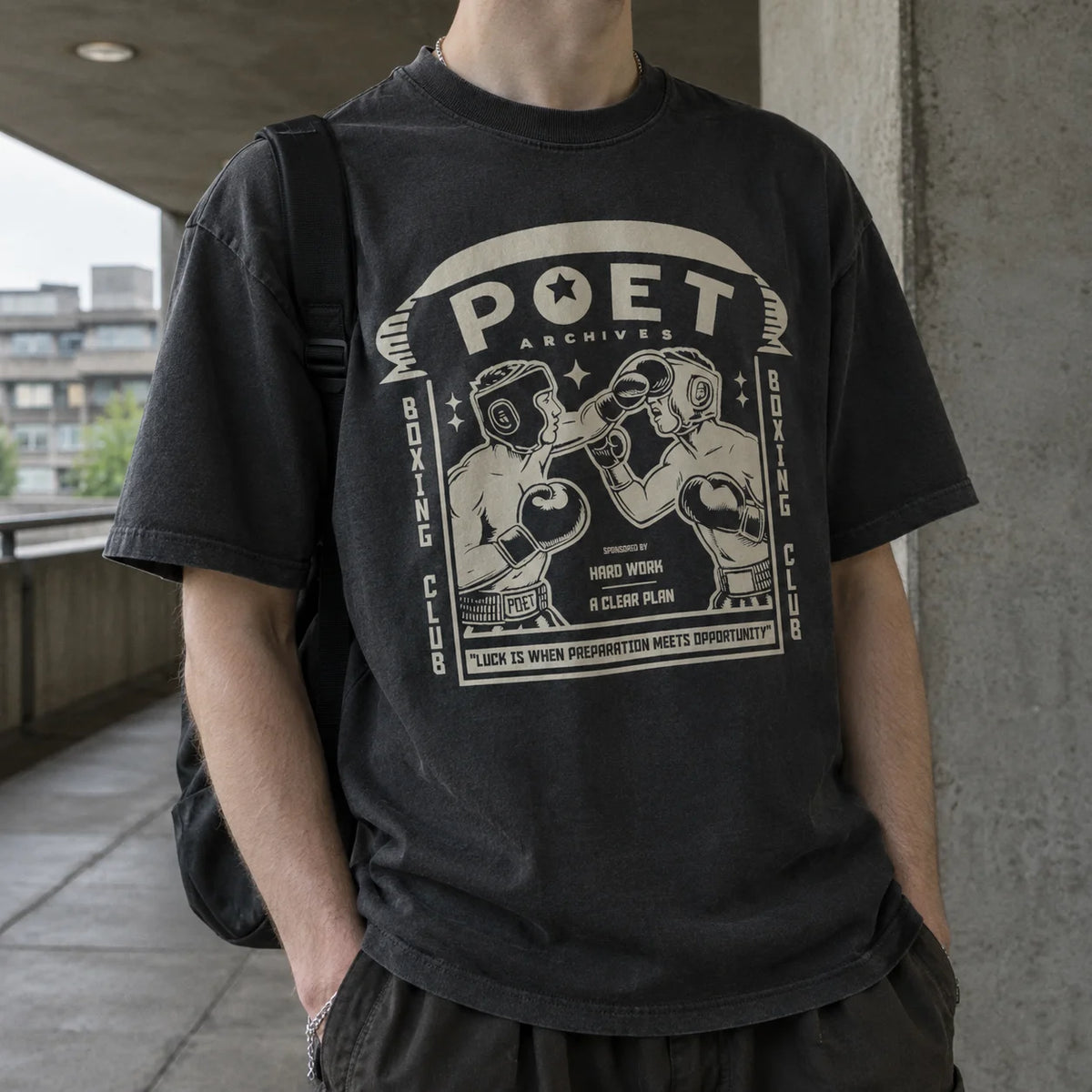 A person wearing a faded black graphic t-shirt with a vintage boxing club design, standing on a concrete walkway.