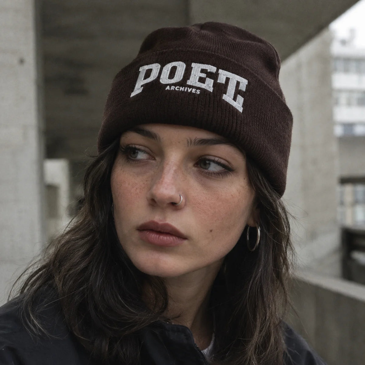 A woman wearing a dark brown ribbed knit beanie with 'POET ARCHIVES' embroidered in white on the front, photographed in an urban concrete setting.