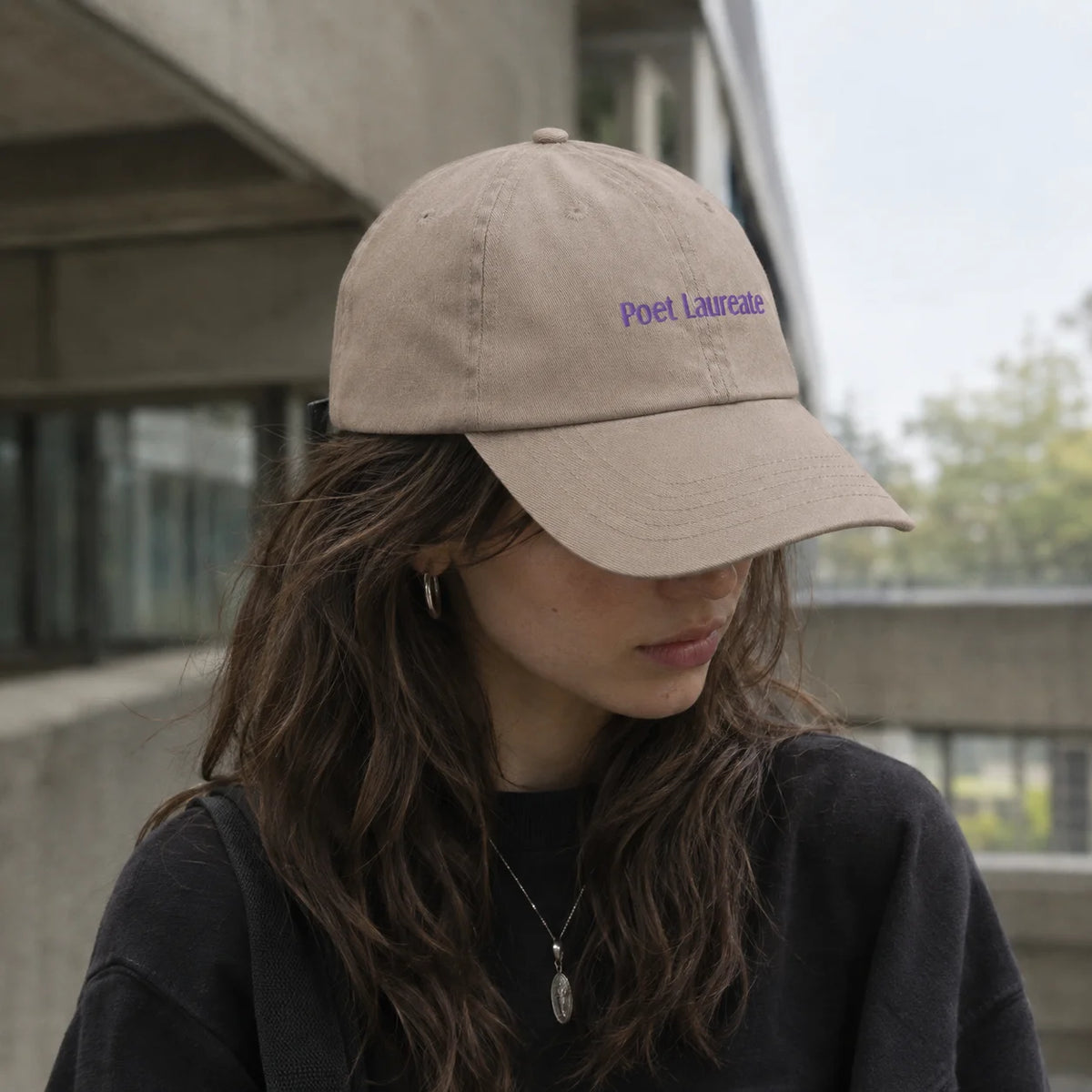 A woman wearing a beige baseball cap with purple embroidered text reading 'Poet Laureate' on the front, photographed outdoors against a modern concrete building.