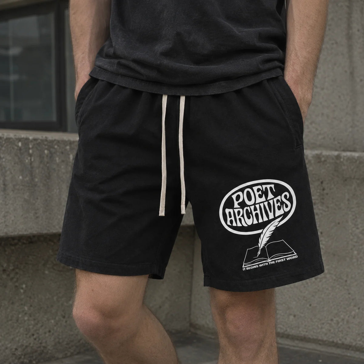 A person wearing black drawstring shorts with a white 'POET ARCHIVES' graphic on the left leg, standing against a concrete wall.