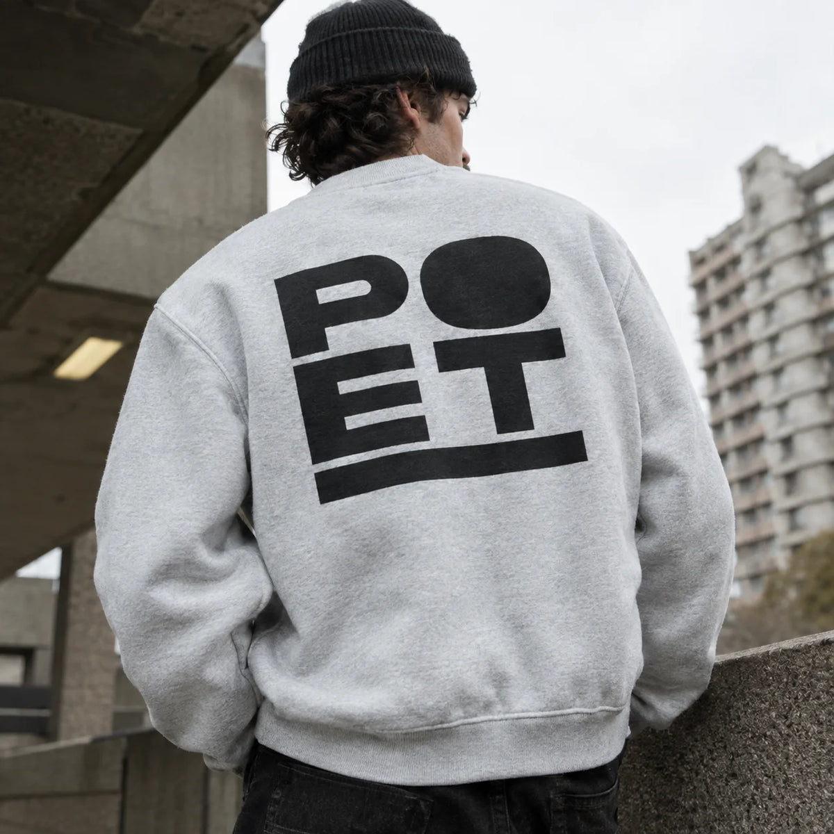 A person viewed from behind wearing a light gray crewneck sweatshirt with a large black 'POET' graphic on the back, paired with a black beanie in an outdoor urban setting.
