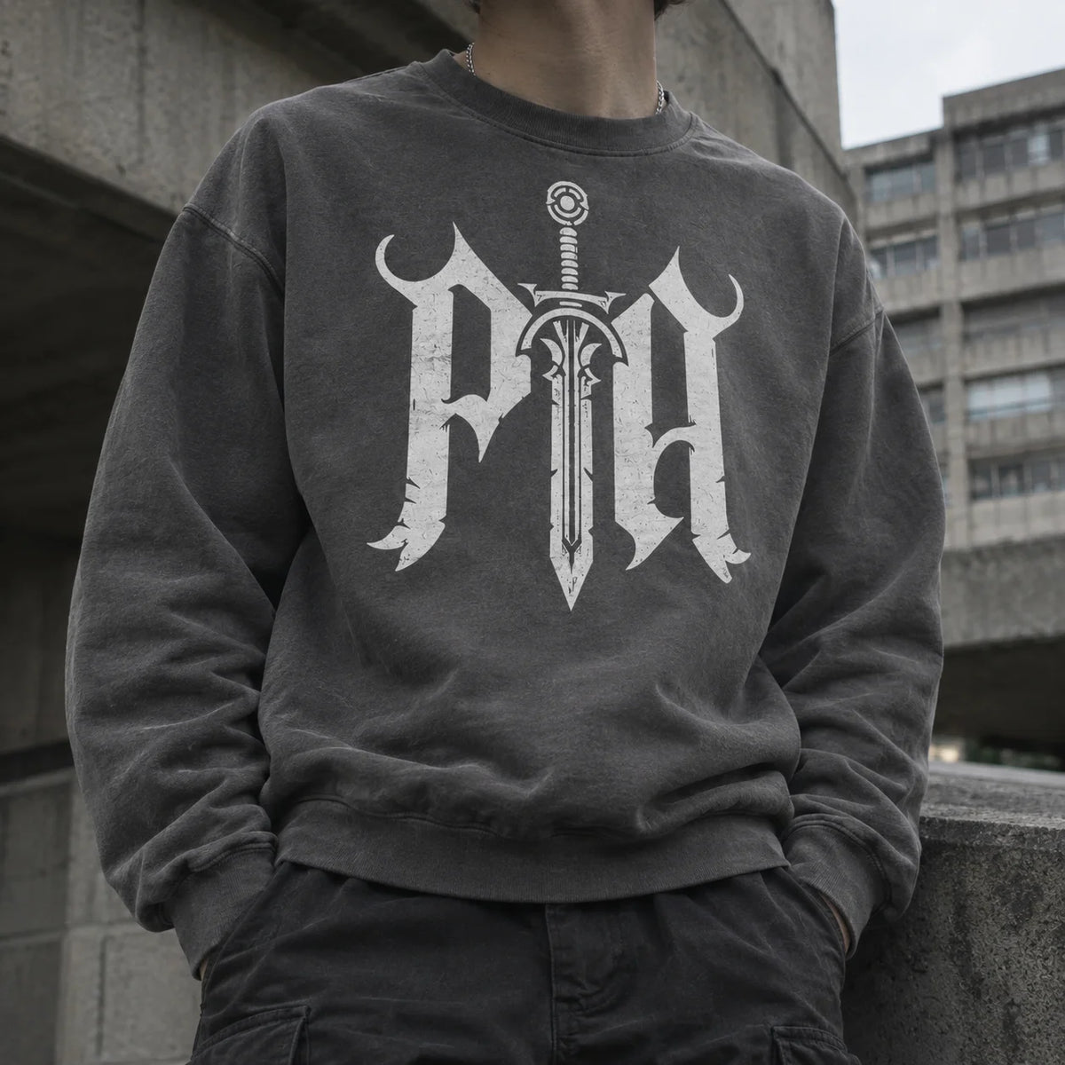 A person wearing a dark charcoal gray crewneck sweatshirt with a large white gothic letter 'P' and sword graphic on the front, paired with dark cargo pants, set against a concrete architectural background.