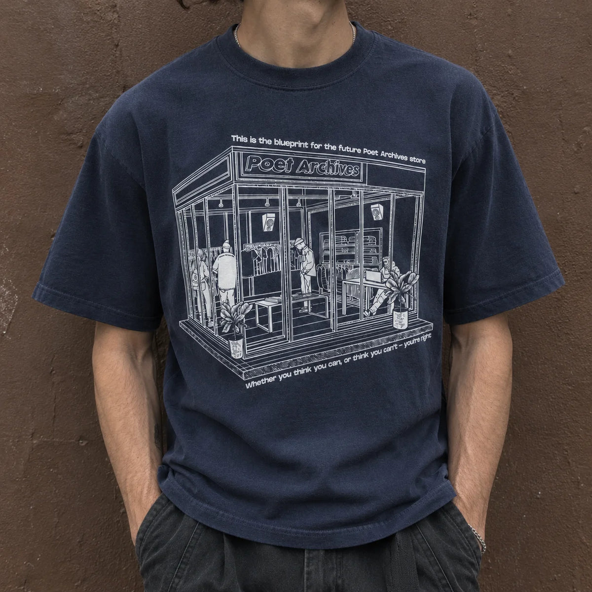 A person wearing a navy blue graphic t-shirt with a white line art print of a clothing store named 'POET Archives'. The background is a textured brown wall.