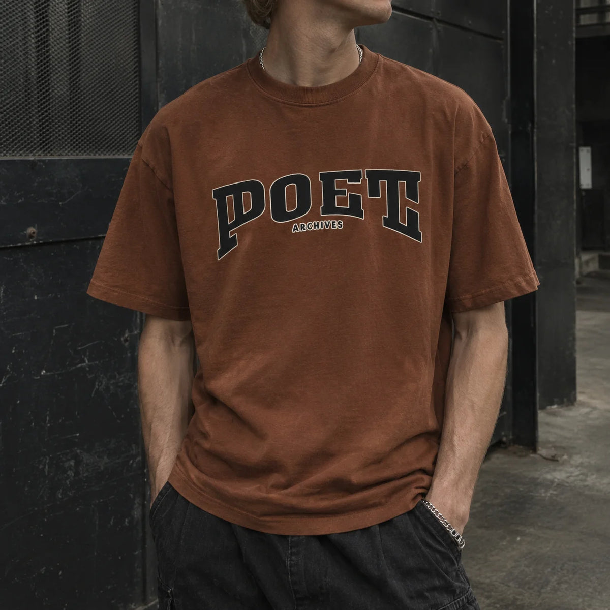 A person wearing a washed brown t-shirt with 'POET ARCHIVES' printed in navy and white arched text across the chest, standing against a dark textured wall.