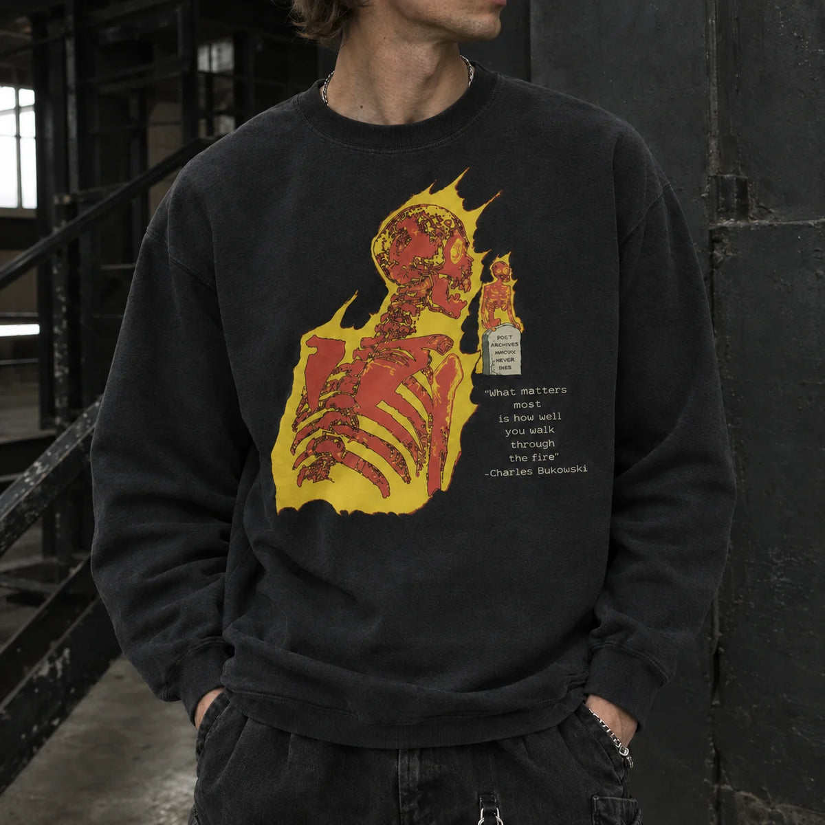 A person wearing a black crewneck sweatshirt with a large red and yellow graphic of a skeleton and text on the front, standing in a dark industrial setting.