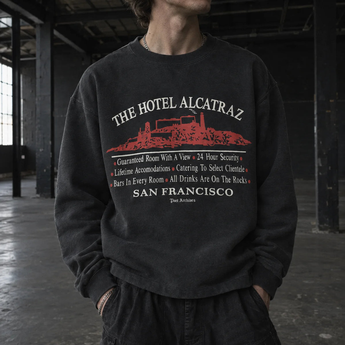 A person wearing a dark charcoal crewneck sweatshirt with a red and white graphic print that reads 'THE HOTEL ALCATRAZ' and 'SAN FRANCISCO' with a red illustration of the Alcatraz prison, photographed in a dark industrial loft setting.