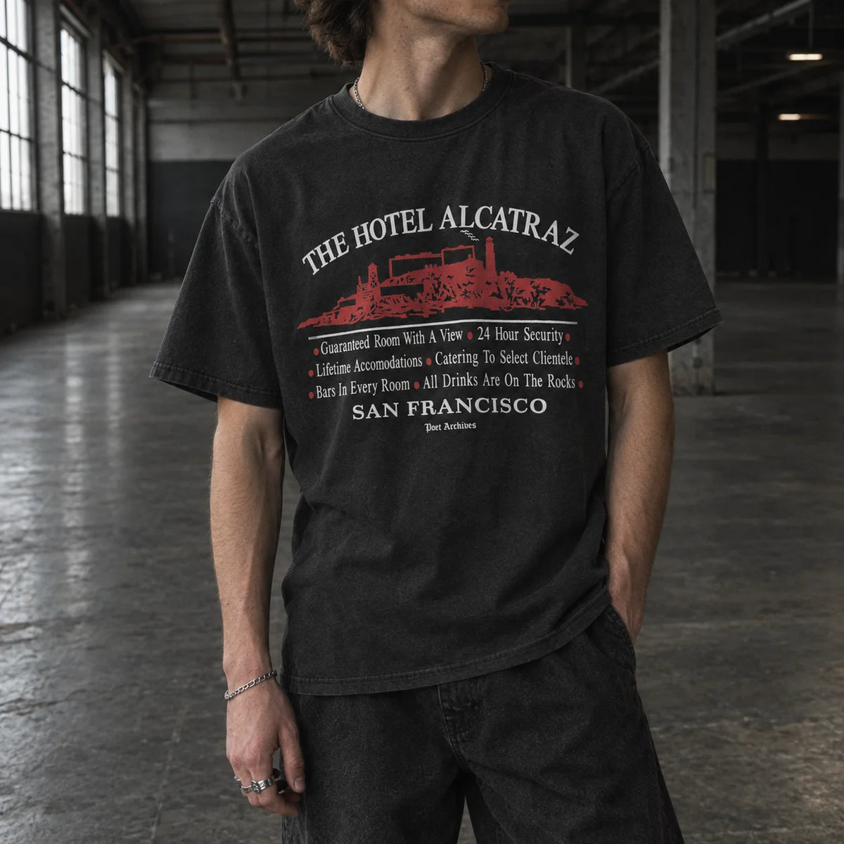 A man wearing a black graphic t-shirt with a red and white print of 'The Hotel Alcatraz' in San Francisco, standing in a large industrial loft with concrete floors and windows.
