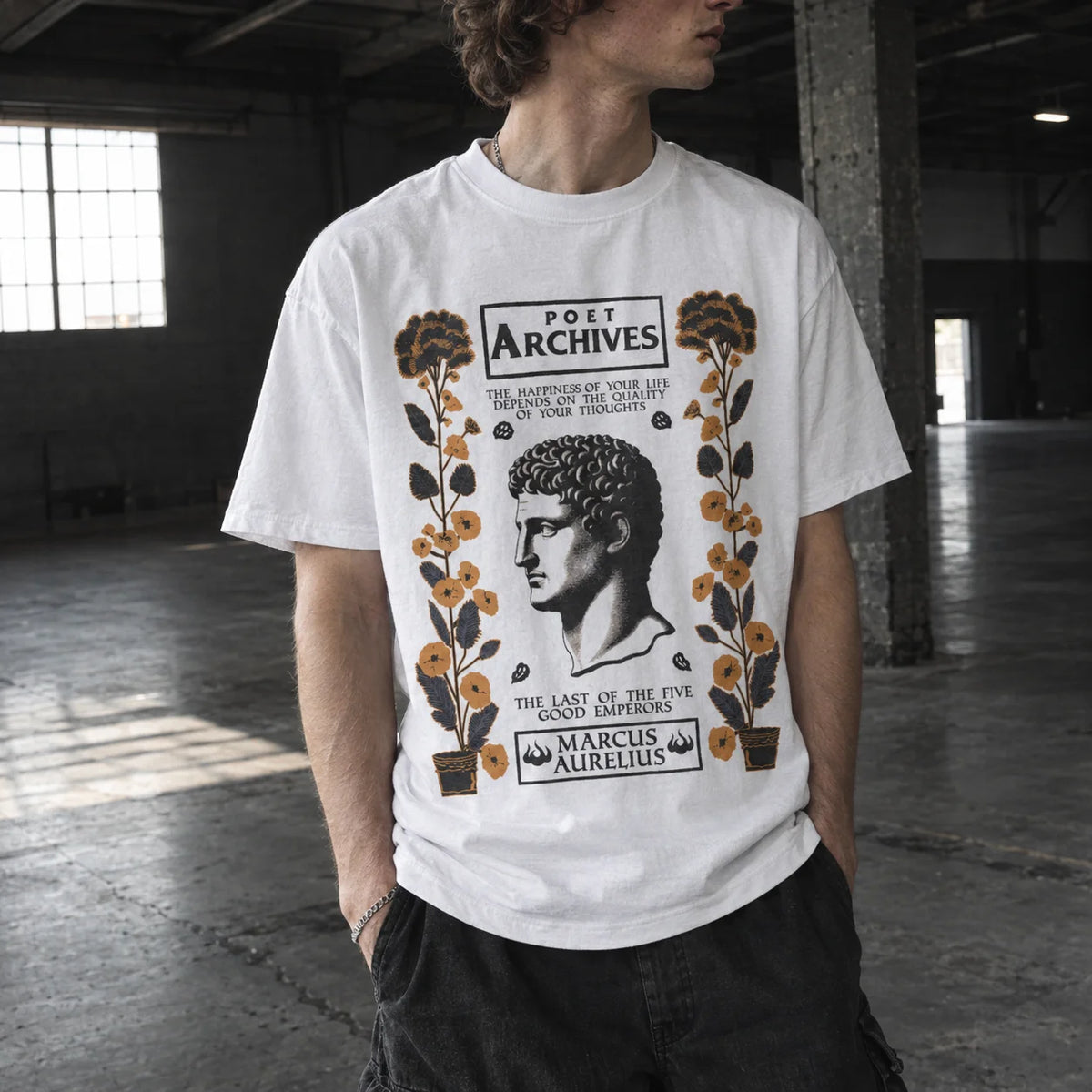 A man wearing a white graphic t-shirt with a vintage-style classical poster design featuring a portrait of Marcus Aurelius and the text 'Poet Archives'. The setting is a dimly lit industrial loft with concrete floors and pillars.