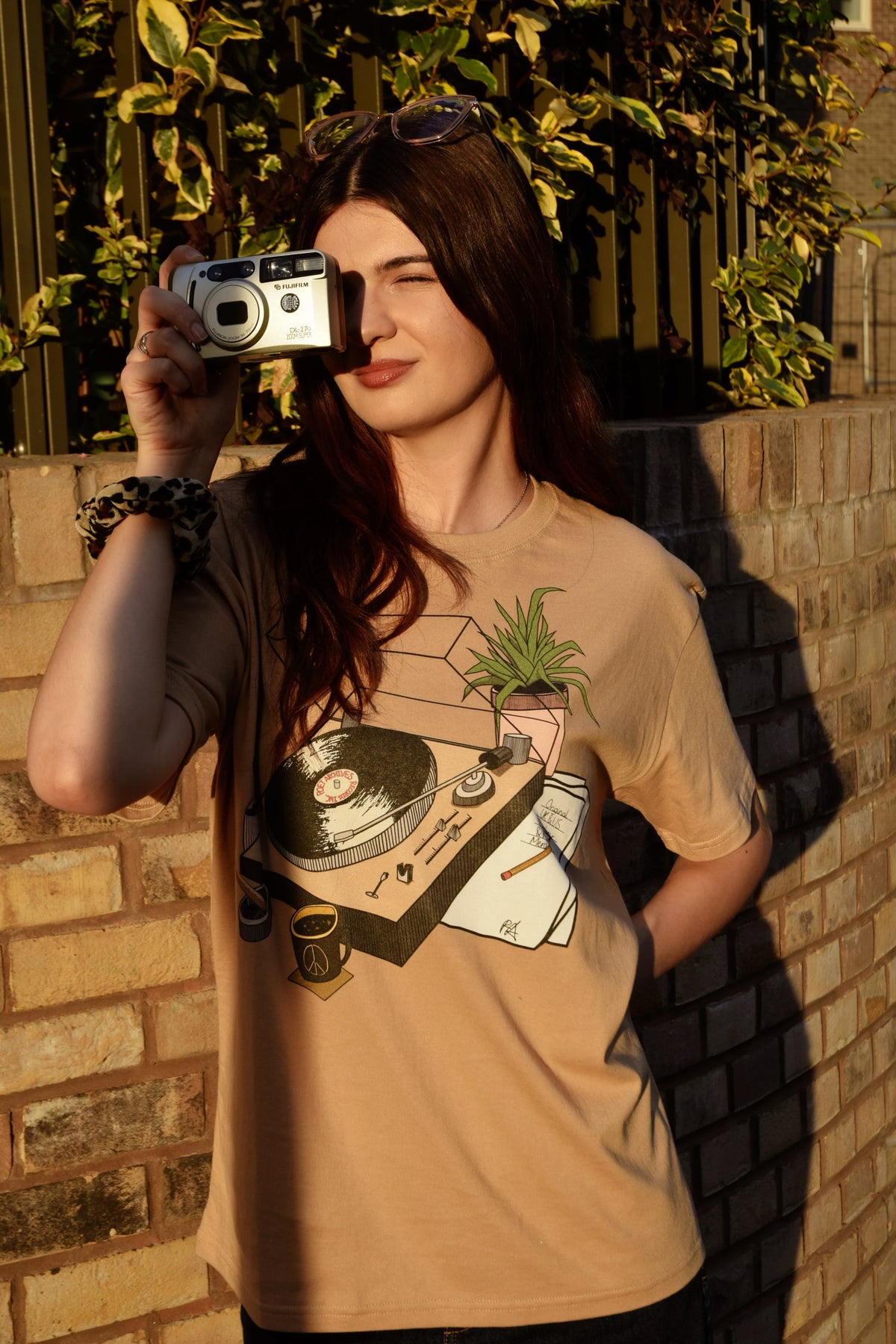 A woman wearing a beige graphic t-shirt with a vinyl record and plant design, holding a silver camera in front of her face, standing against a brick wall with green foliage in the background.