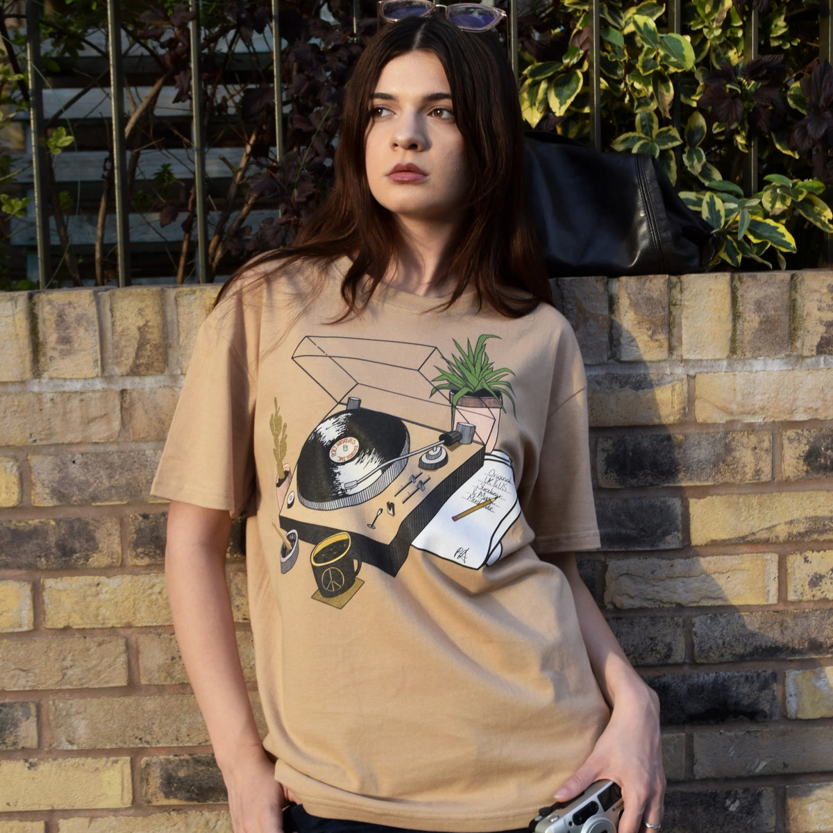 A woman wearing a beige graphic t-shirt with a vinyl record and plant design, paired with dark blue jeans, standing against a brick wall with green foliage in the background.