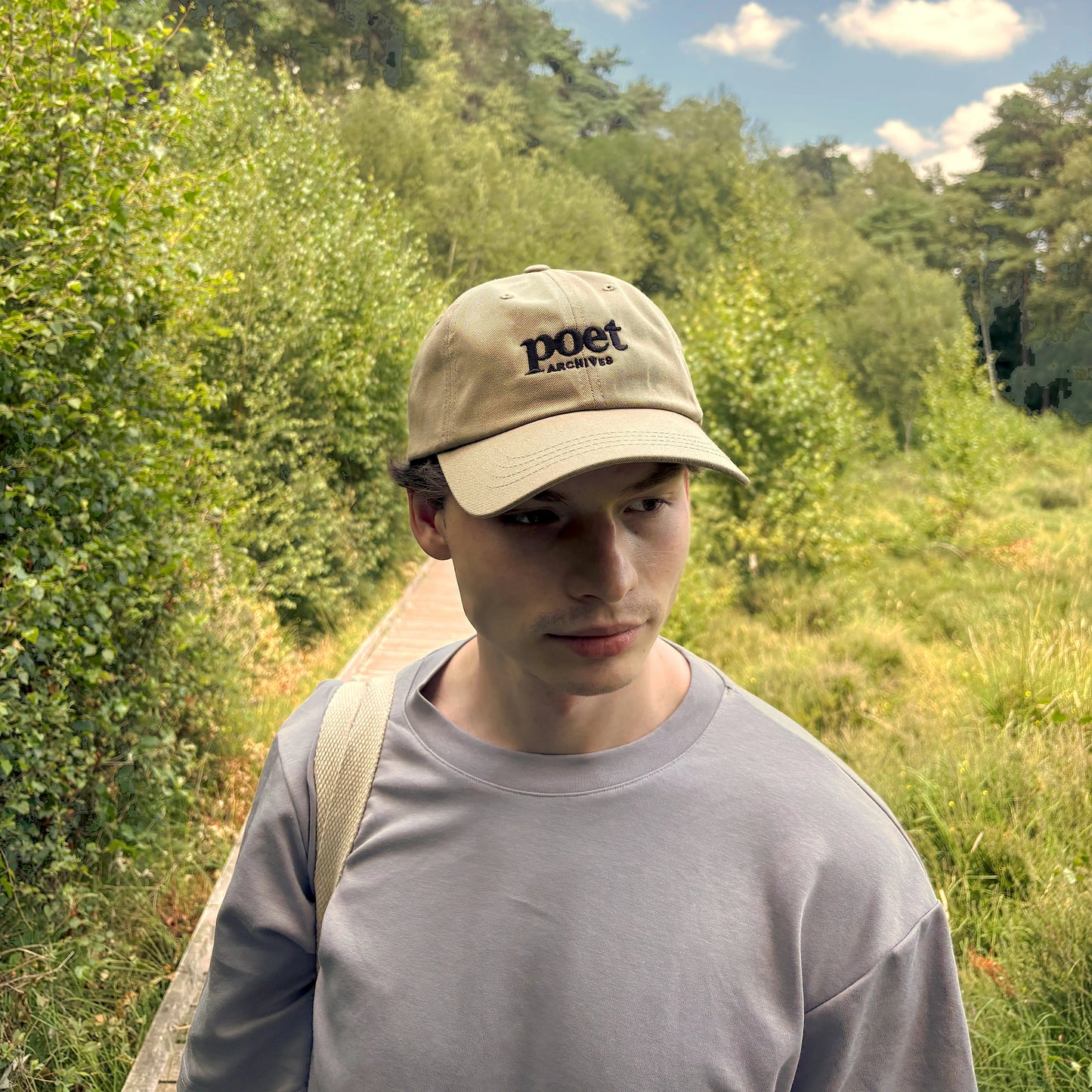 A man wearing a beige baseball cap with 'poet' embroidered on the front, standing outdoors on a path surrounded by green trees and grass under a blue sky.