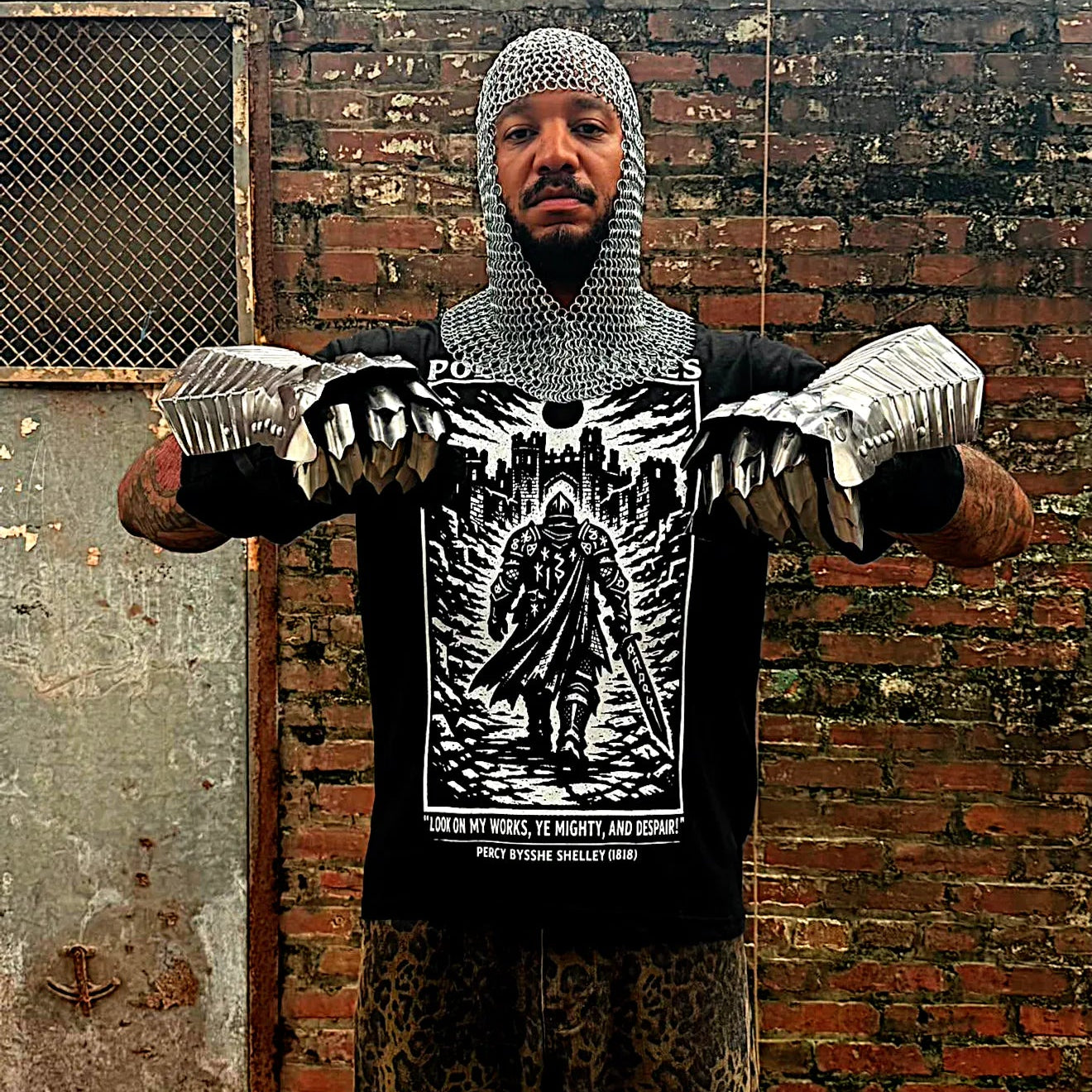 A man wearing a silver chainmail coif, silver clawed finger gloves, and a black graphic t-shirt stands in front of a brick wall. The graphic on the shirt features a knight in armor with the text 'THE KING IS NOT DEAD'.