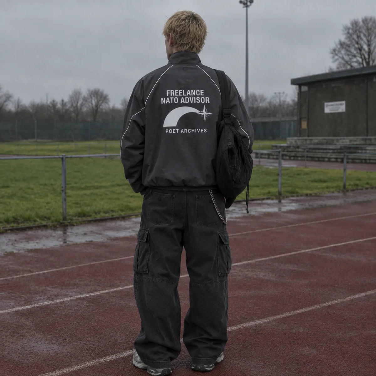 A man stands on a wet red running track wearing a black jacket with a large white back graphic that reads 'FREELANCE NATO ADVISOR' and 'POET ARCHIVES', paired with black cargo pants, photographed from behind in an outdoor overcast setting.
