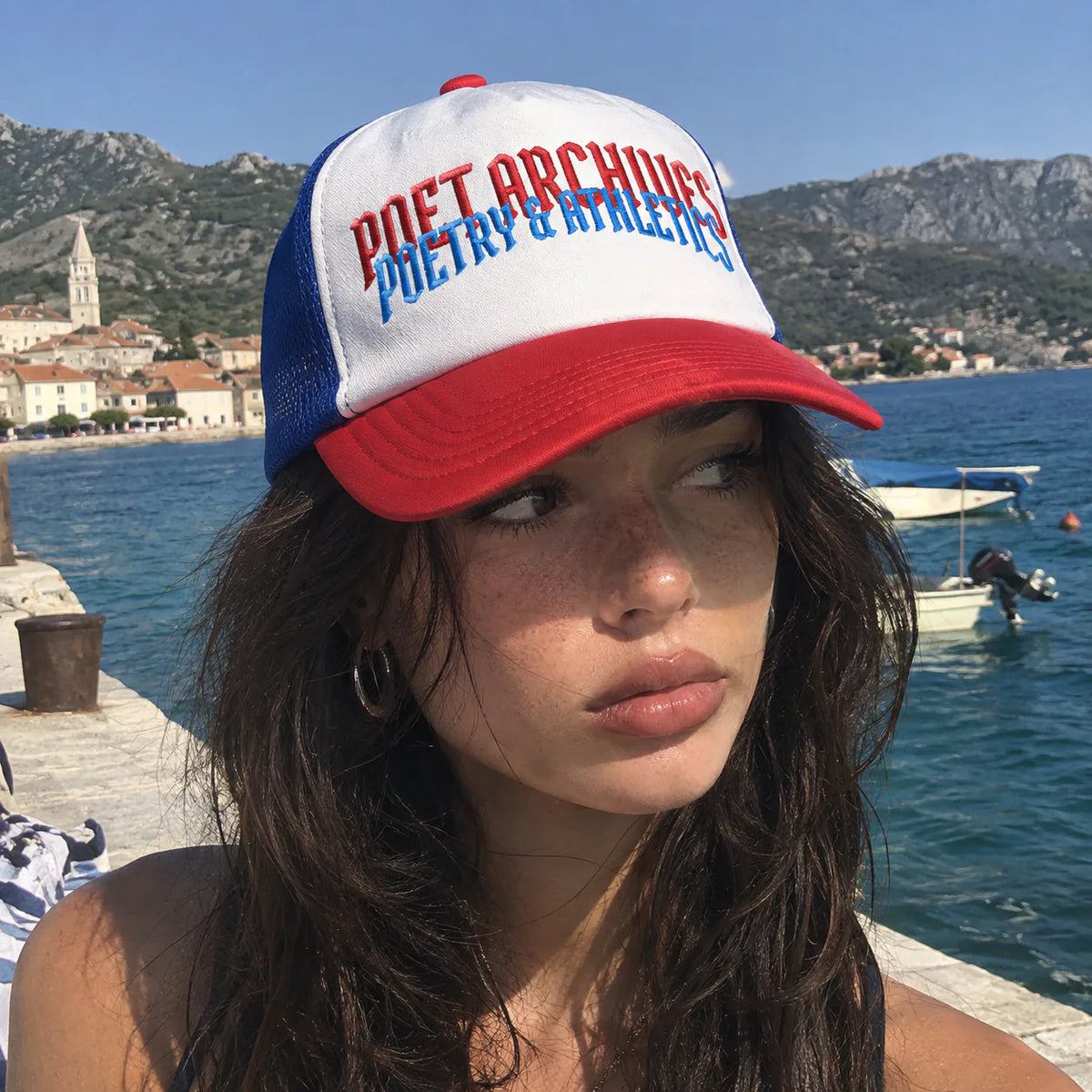 A woman wearing a red, white, and blue trucker hat with the text 'POET ARCHIVES POETRY & ATHLETICS' in front of a coastal harbor background.