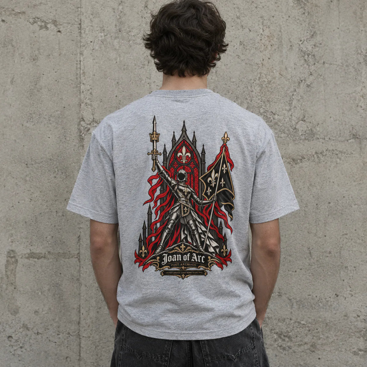 A person viewed from behind wearing a light gray heather t-shirt with a large graphic of a gothic knight and the text 'Joan of Arc' on the back, standing against a textured concrete wall.