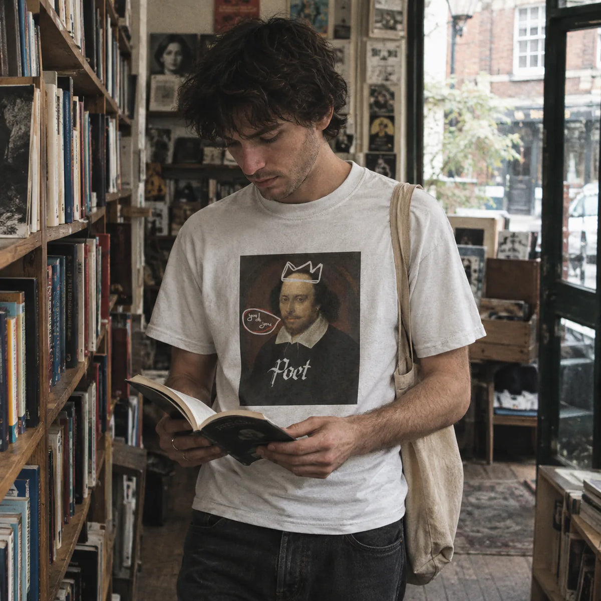 A man in a bookshop wearing a white graphic t-shirt with a Shakespeare print and a canvas shoulder bag, reading a book.