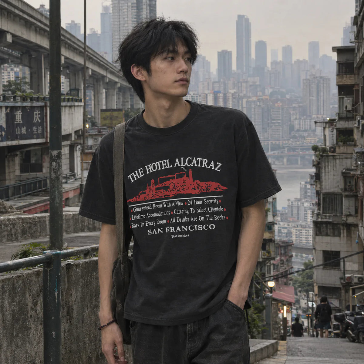 A man wearing a black graphic t-shirt with a red and white 'The Hotel Alcatraz' print, standing on an outdoor urban staircase with a city skyline in the background.