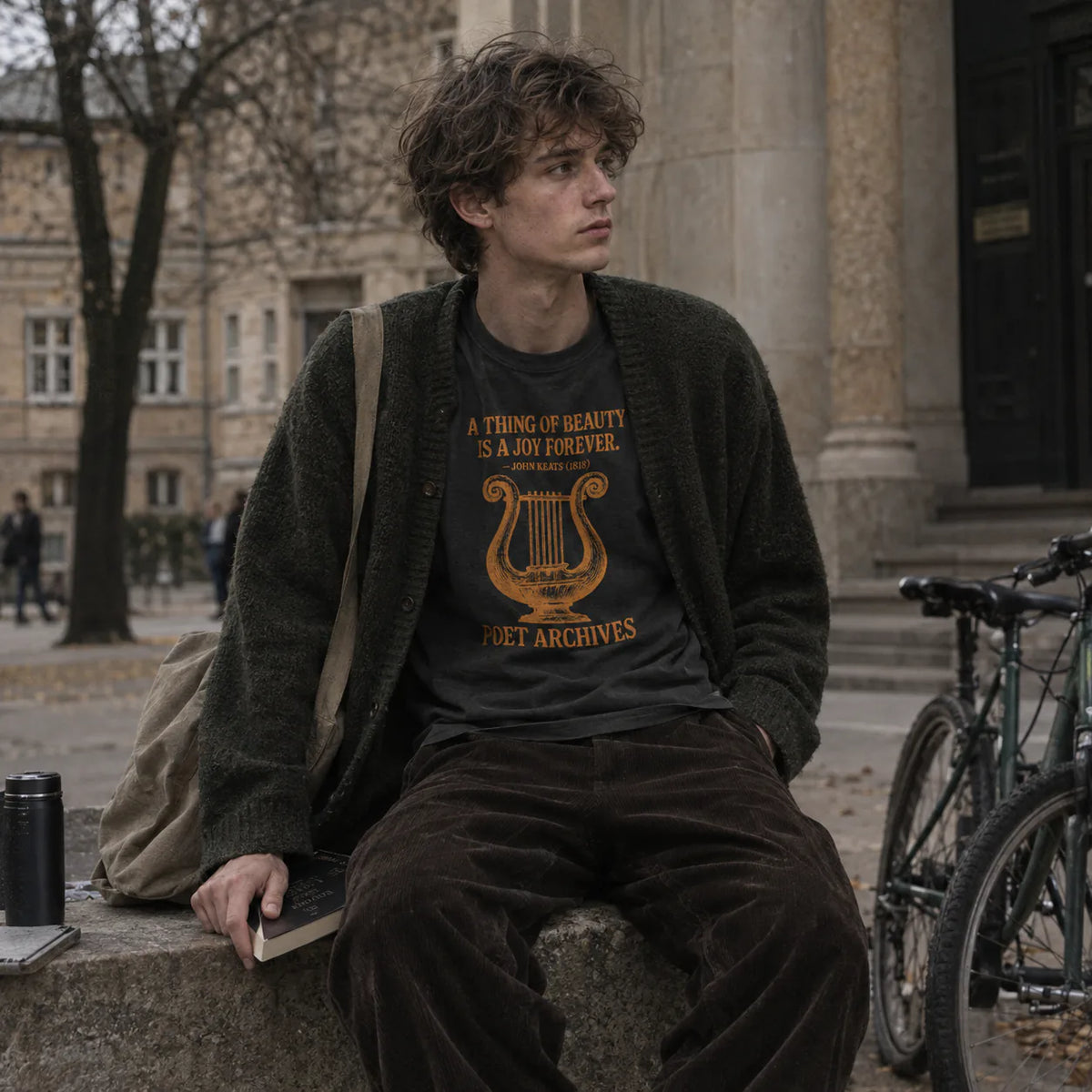 A young man sits on a stone bench in an outdoor urban setting, wearing a dark brown knit cardigan over a black graphic t-shirt with a gold harp and the text 'A THING OF BEAUTY IS A JOY FOREVER. POET ARCHIVES'.