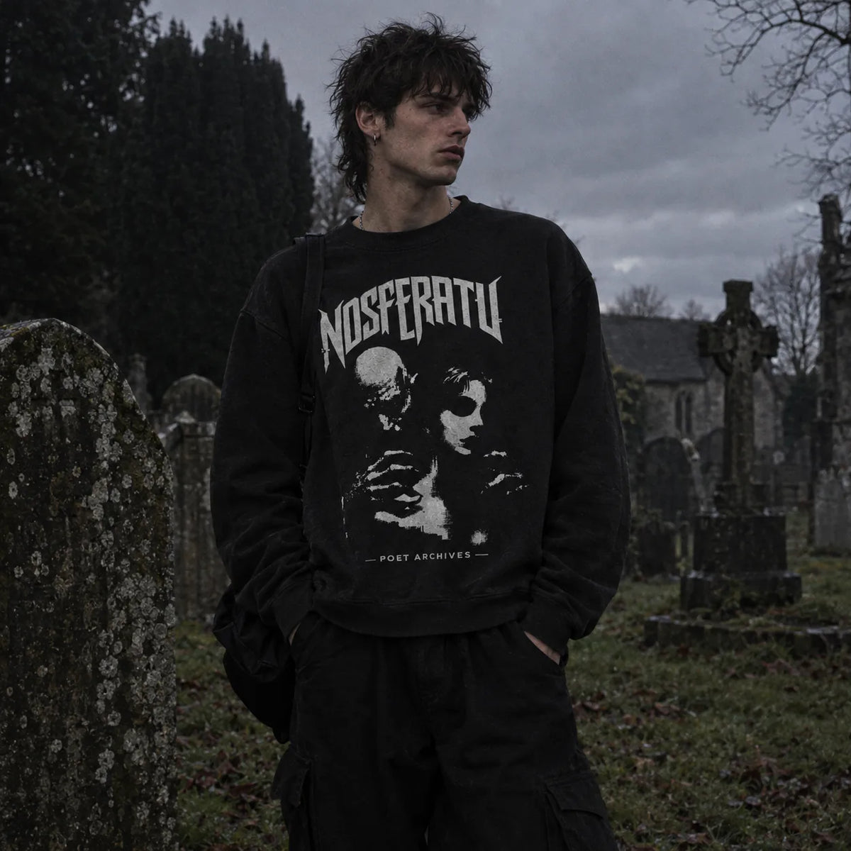 A man standing in a cemetery wearing a black crewneck sweatshirt with a white graphic print reading 'NOSFERATU' and 'POET ARCHIVES'.