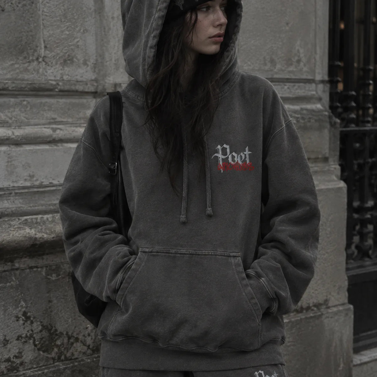 A person wearing a faded dark gray hoodie with a small white and red embroidered chest graphic, standing outdoors against a stone wall.
