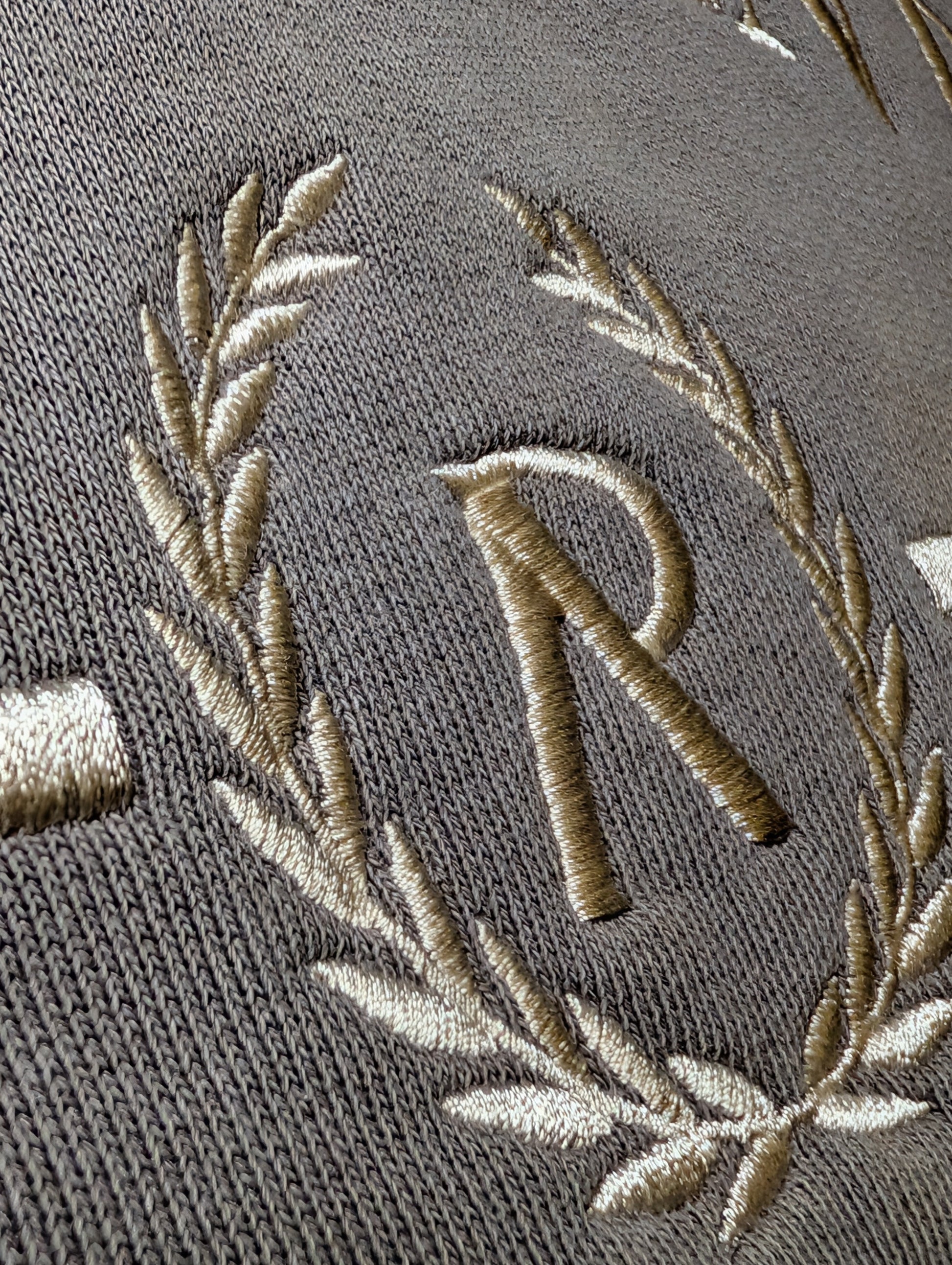 Embroidered design with a letter 'R' and laurel leaves on a textured surface.