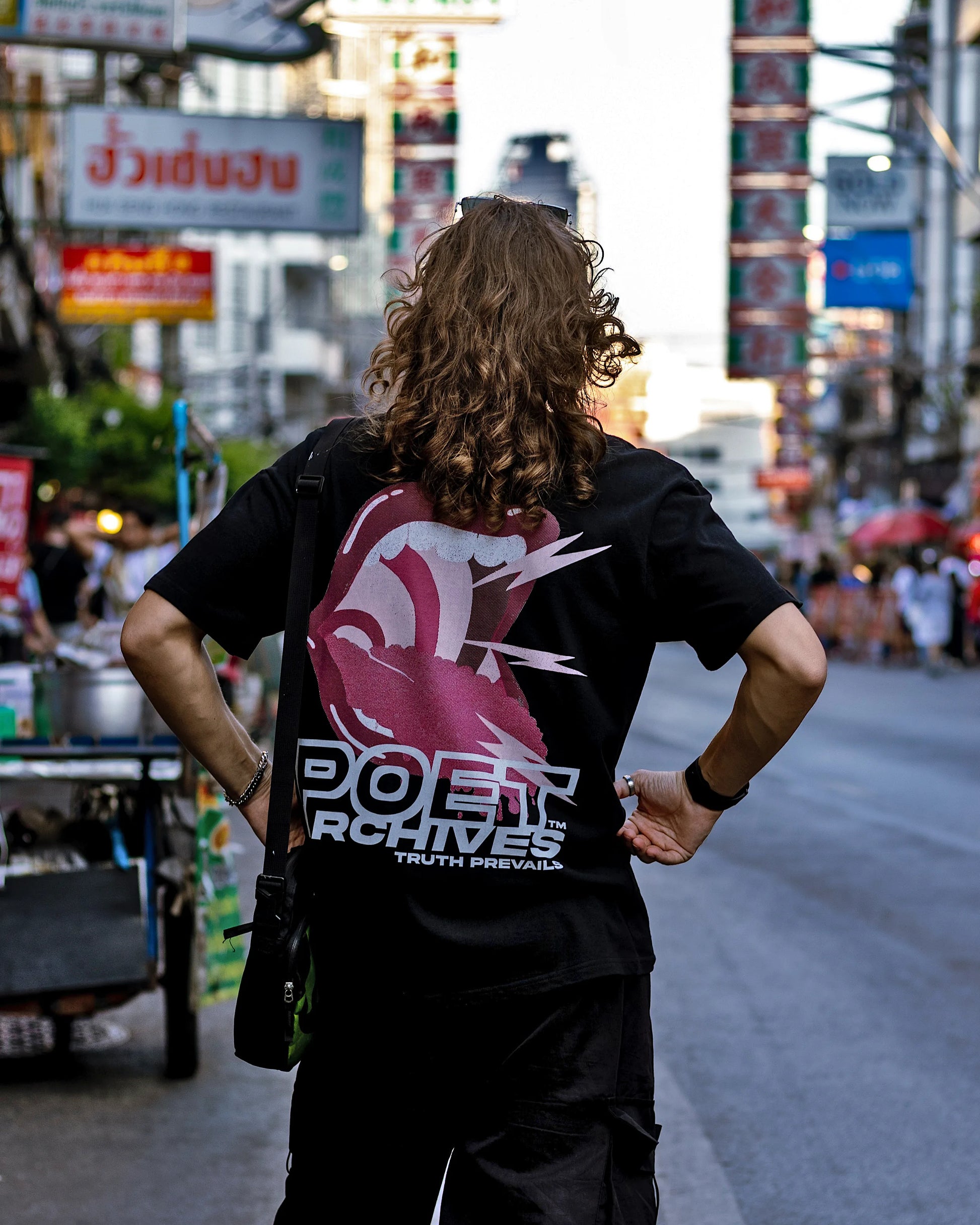 Person wearing a black t-shirt with a logo and text on the back, standing on a street with blurred background