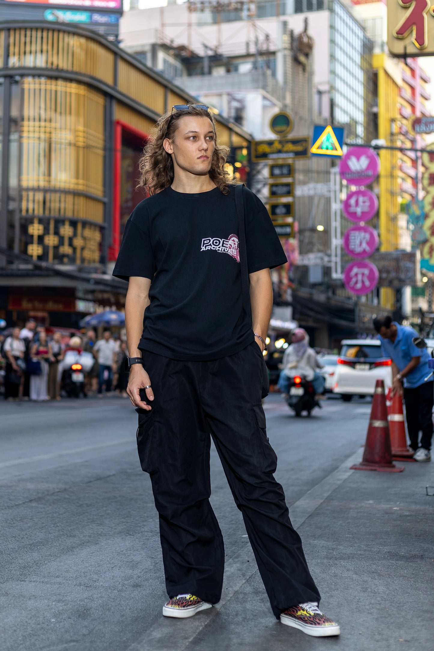 Person wearing a black t-shirt with a logo and dark pants standing on a busy street with neon signs in the background.
